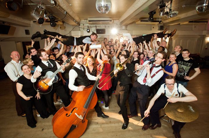 Band members surrounded by dancers, with piano player and trumpet player crowd surfing